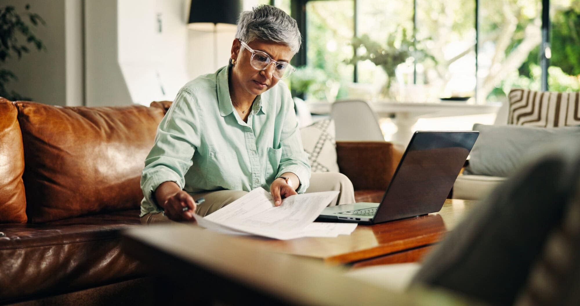 Mature woman, remote work and document for finance, budget review or asset management. Reading, freelancer and person with paper in living room for accounting, insurance or expenses on laptop in home.